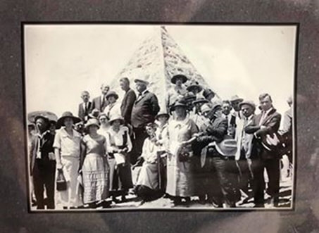 A photograph of a group of people dressed in 1920’s clothing gathered in front of a stone pyramid about 12 feet tall.