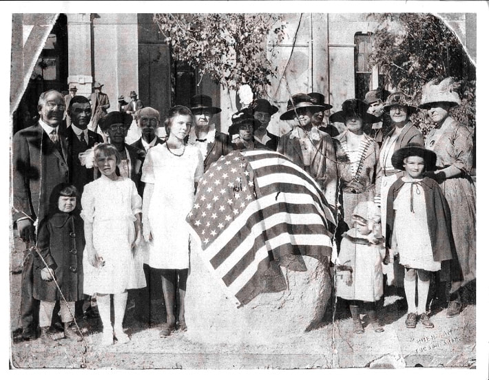 A group of adults and children dressed in 1920’s clothing gathered around a stone monolith about four feet high. The stone is covered with a 48-star U.S. flag. The women and two of the children are wearing hats.