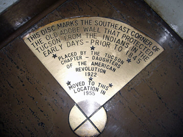 A brass disc embedded in a corner of linoleum flooring with a quarter-circle label that reads “This disc marks the southeast corner of the Old Adobe wall that protected Tucson from the Indians in the early days - prior to 1845 / Placed by the Tucson Chapter - Daughters of the American Revolution 1922 / Moved to this location in 1955”