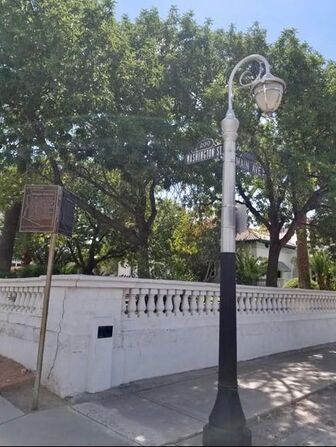 A corner with a low wall with balustrade along a sidewalk. Street signs read “200 Washington St” and “Main Ave.” A rectangle that appears to be a plaque is mounted on the wall about 30-inches from the ground.