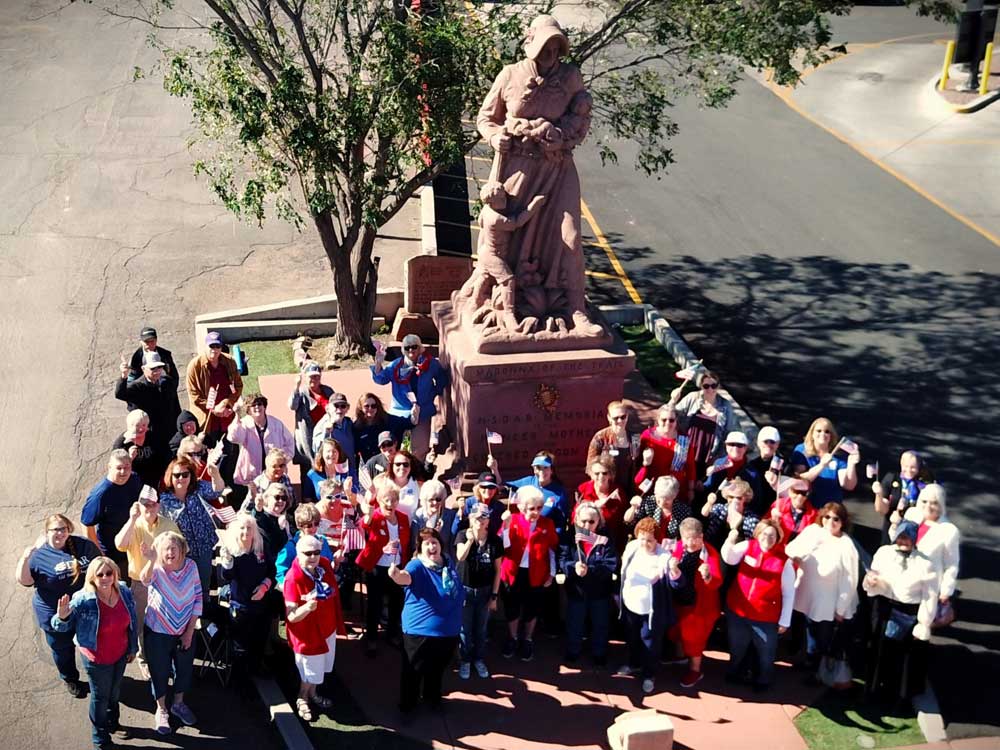 An aerial photograph of the Madonna of the Trail statue with a group of about fifty people gathered in front and smiling at the camera.