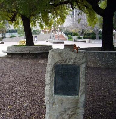 An outdoor scene of a park or plaza with two trees in mid-ground. A stone monolith with a marker with unclear writing is in the foreground. Former Spanish Presidio.