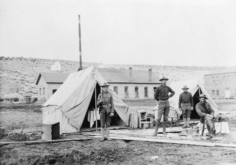 A vintage photograph of four men in front of two tents in a desert scene. A flagpole and two buildings are in the background.