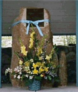 A snapshot of a standing stone with plaque, sporting a large blue ribbon. A large spray of flowers stands in front of the marker. Celebrates Sharlot Hall.