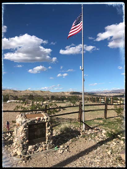 A snapshot of the marker in the foreground, next to a flagpole, the cemetery in mid-ground, and mountains in the background.