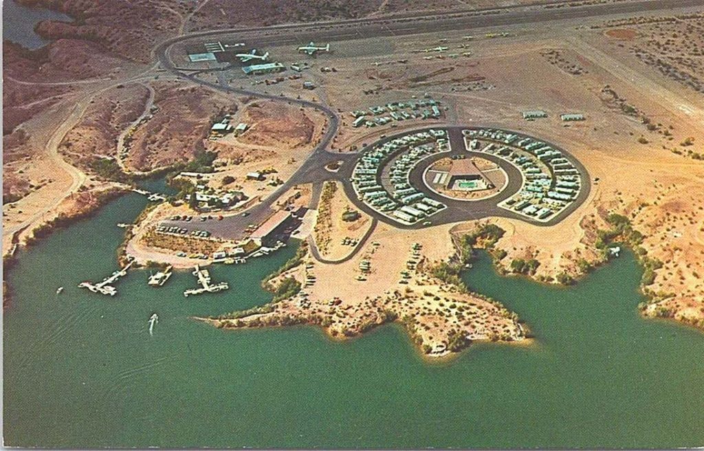 An aerial photograph of a desert area next to a river, showing a runway and two planes in the background. A circular blacktop area has about sixty one-story buildings arranged in a neat, orderly manner.