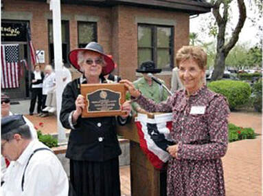 A snapshot of adults in period costume in front of the school. Two women are holding the marker for the little red schoolhouse.