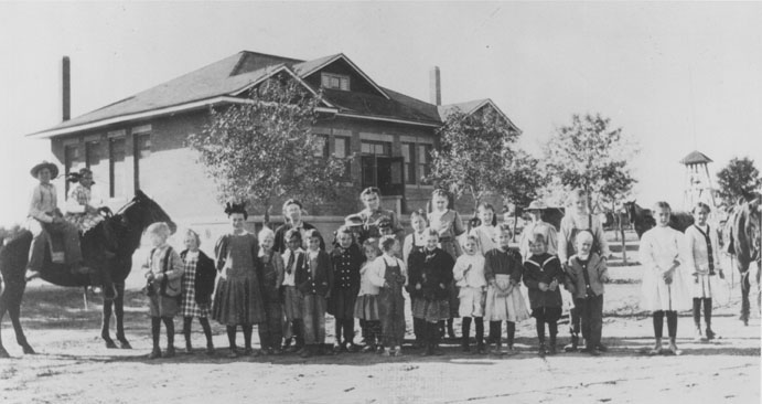 A photograph of a one-story brick schoolhouse with about twenty children and their teachers standing in a group. Two children are mounted on horses or mules.