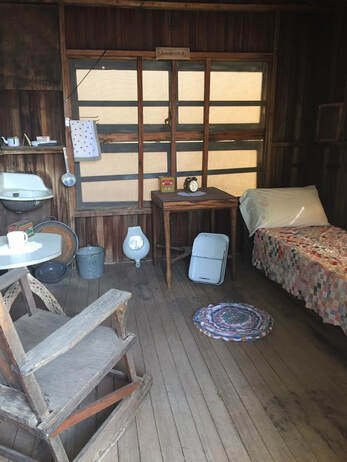 Snapshot of the interior with a bed, small rag rug, basin attached to the wall, rocking chair, small table, and other items.