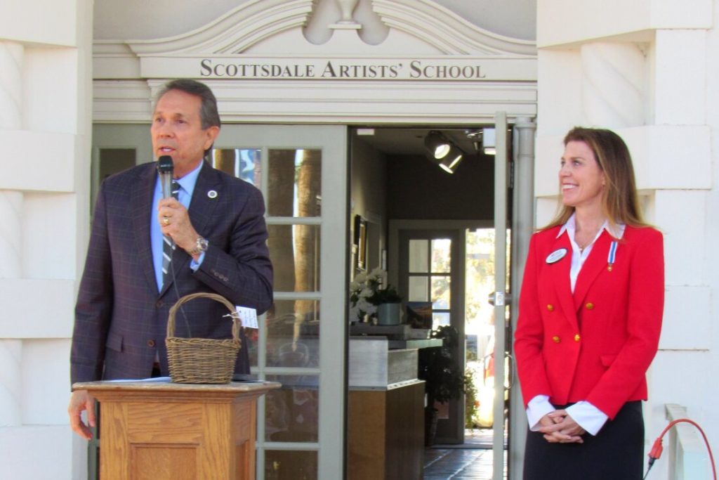 Mayor David Ortiz is speaking from a podium with Karen Andrews standing nearby. The open doors to the school building are shown behind them, giving a glimpse into the historic building.