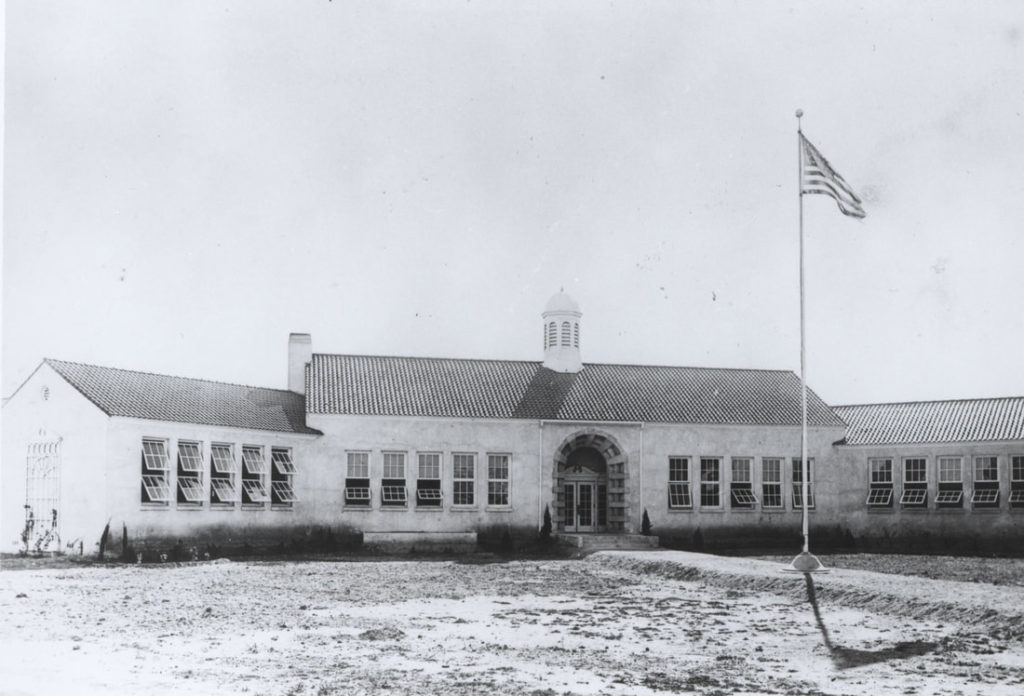 A vintage photograph of a tall one-story school building with a central wing with two groups of five windows and two flanking wings, each with five windows. The courtyard may be bare soil. A walkway to the front door contains a flagpole. Scottsdale Grammar School 2.