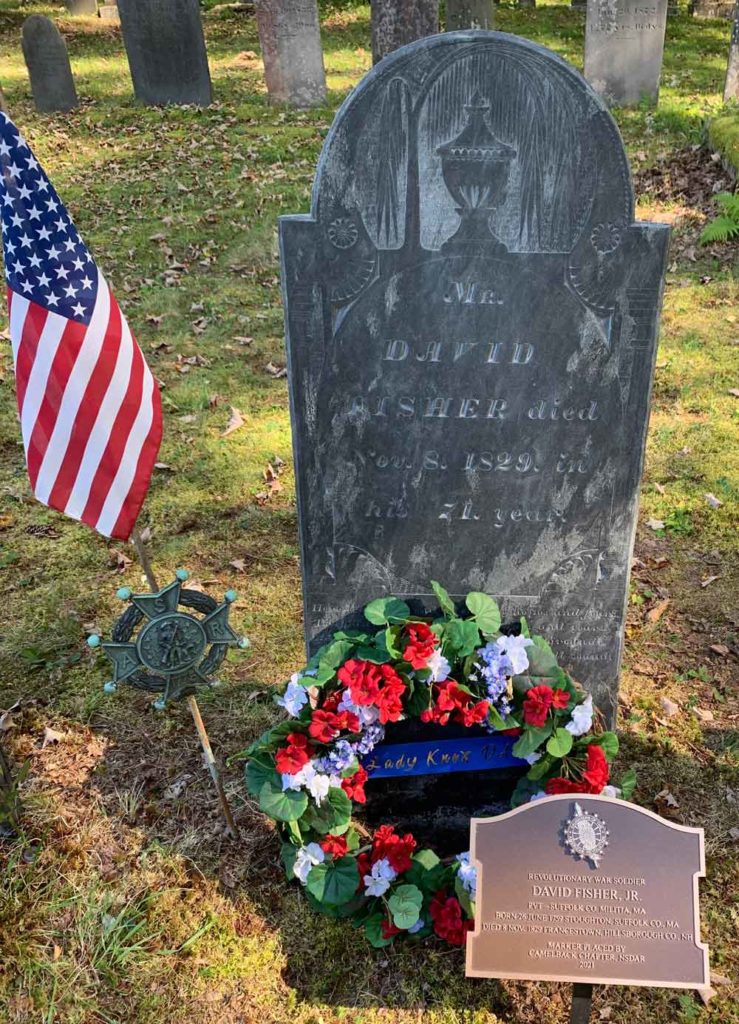 A snapshot of a slate standing gravestone with a marker in front. A small floral wreath and a flag are placed near by.