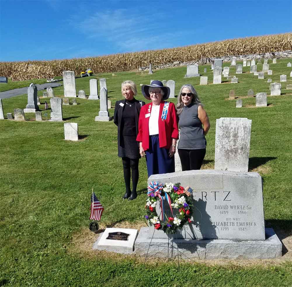 Three women in a grassy, hillside cemetery, standing next to a family memorial, with the marker set beside it.