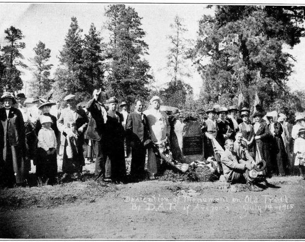 A photograph of a group of people dressed in early 20th century clothing gathered in front of a stone about 5 feet tall with a marker.