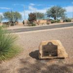 A photo showing the marker embedded onto a boulder and placed on the ground in front of flag poles at the Southern Arizona Veterans’ Memorial Cemetery.  